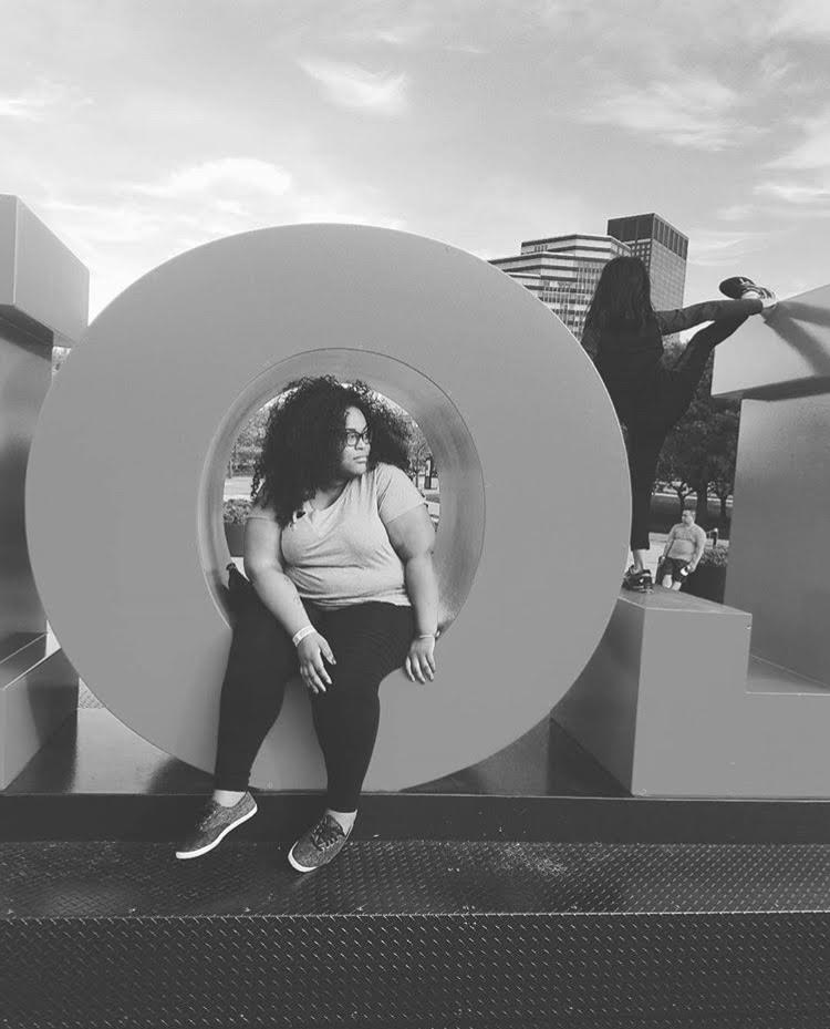Ronnie, a Black perosn, sits in the O in a sign while traveling. The photo is in black and white, and they are wearing sneakers, black leggings and a short sleeve t shirt. 