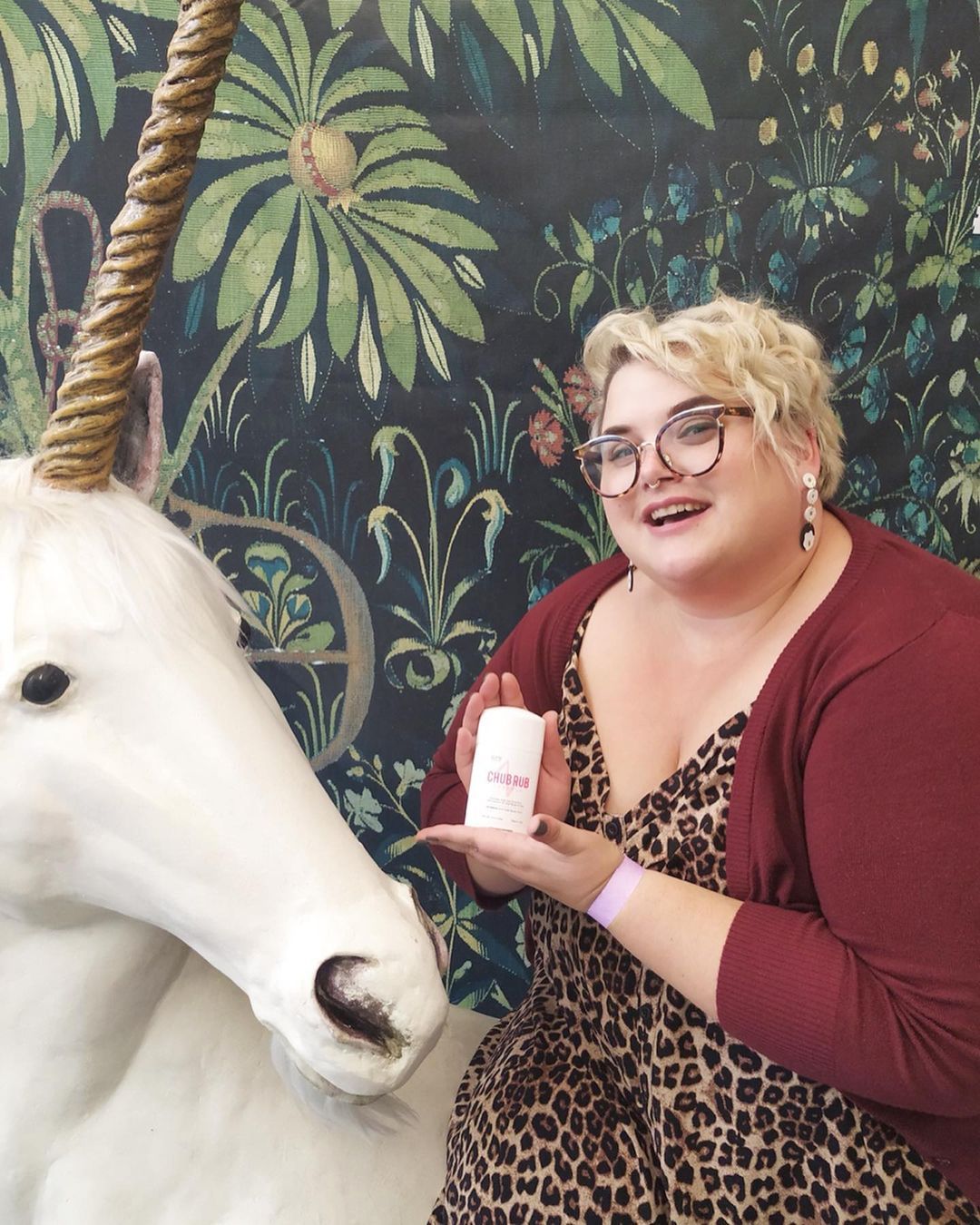 A woman stands in front of a jungle themed background. She is plus sized, with short blonde hair, amd is wearing a leopard print dress and a burgundy cardigan. she is holding a stick of Chub Rub in her hands. Beside her is a white unicorn. Chub Rub Giveaway