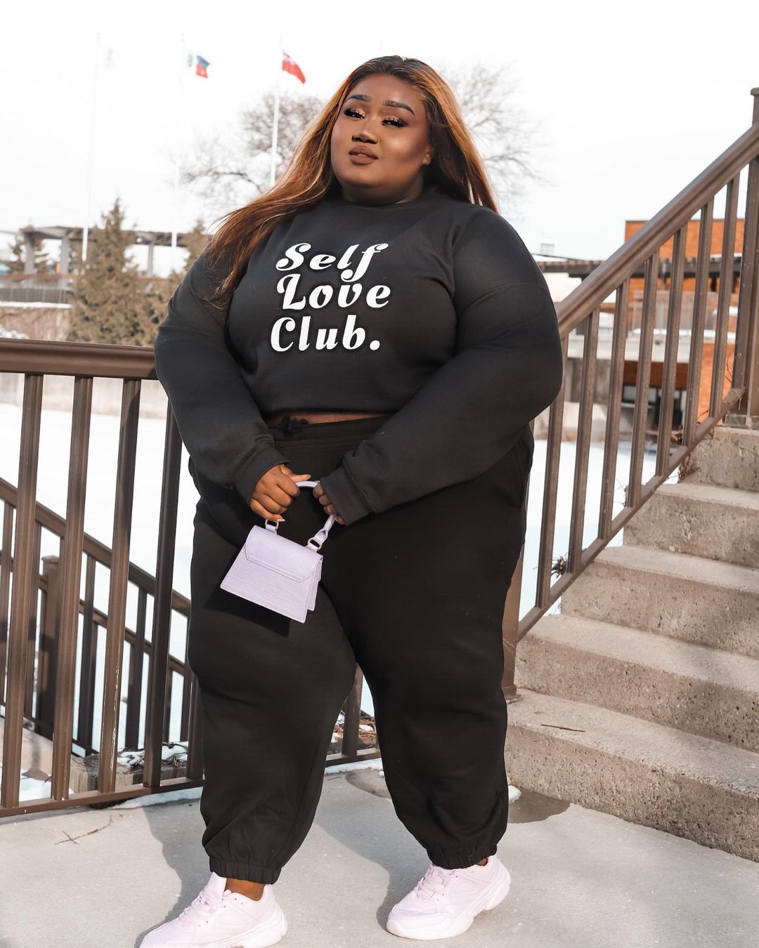 Louange.m posing by a stairway, wearing black sweatpants, a sweatshirt with "Self Love Club" written on the chest. She is holding a white mini bag and wearing white sneakers. Size 24 + model.