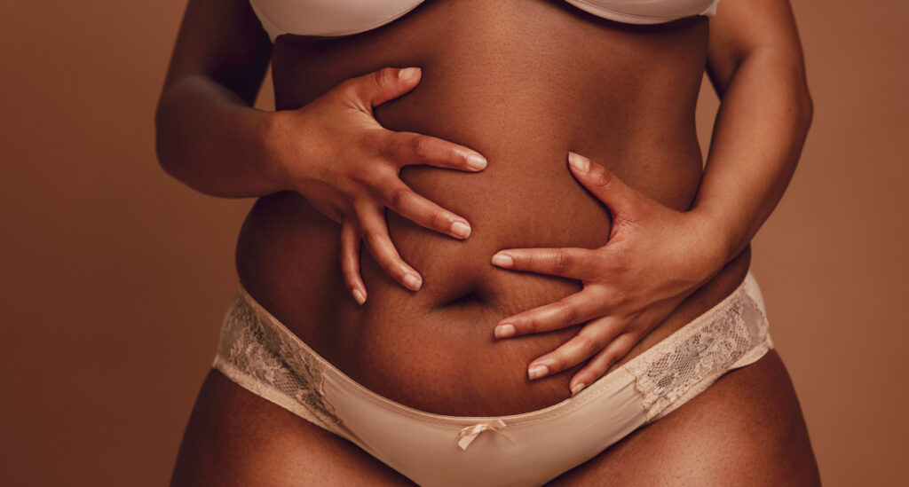 Cropped shot of black female in underwear holding her fat belly. Plus size woman in lingerie holding her belly with both hands on brown background.medical fatphobia during the pandemic