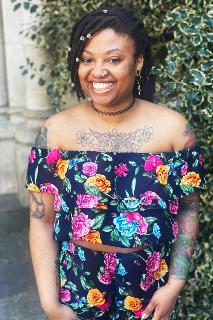 A Black person with tattoos stands in front of a green bush. Their hair is in locs and has some gold bangles throughout them. They are smiling, facing the camera wearing a floral two-piece top. The top has shoulders that come down to the sides, and cuts off right above their belly button. They are wearing a black choker.