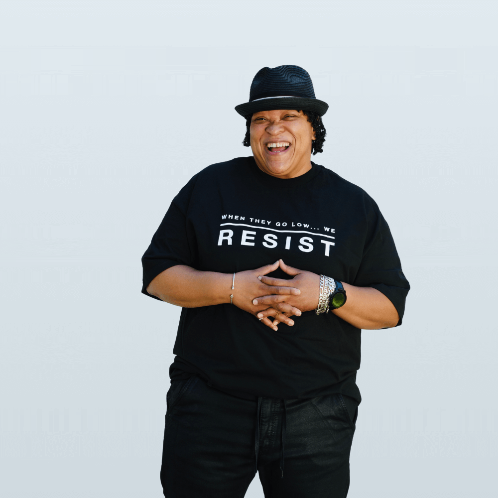 A Black person stands with their hands folded over thei chest. Their har is short, and curly. They are wearing a black fedora and laughing into the camera. They are wearing a short sleeved androgynous black t-shirt that reads "When they go low...we resist" by the Brand Haute Butch, a clothing line dedicated to Androgyny