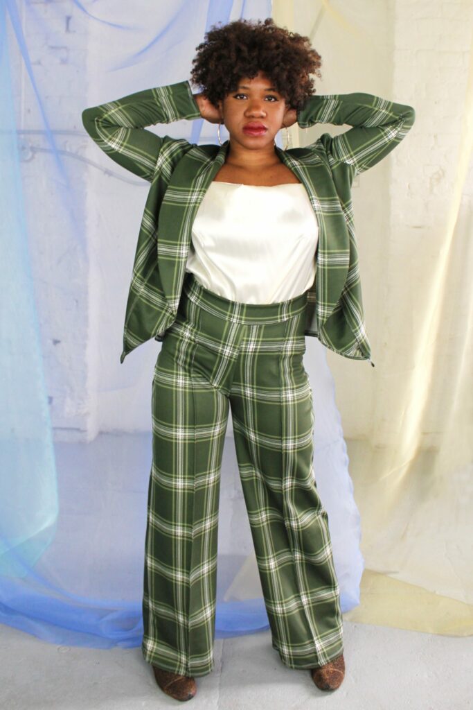 A Black person stands with their arms bent towards their head. Their hair is curly and frames their face. They are wearing a two-piece green plaid suit with a white satin blouse. they are standing in front of a light colored background.