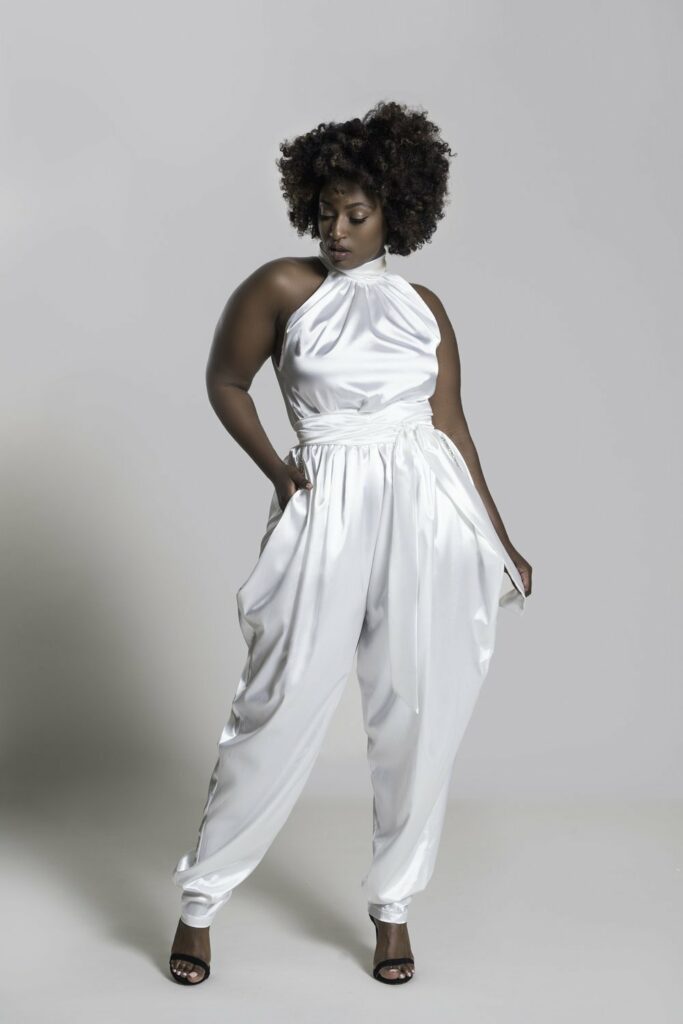 a Black person stands in an off-white satin jumpsuit with no sleeves.