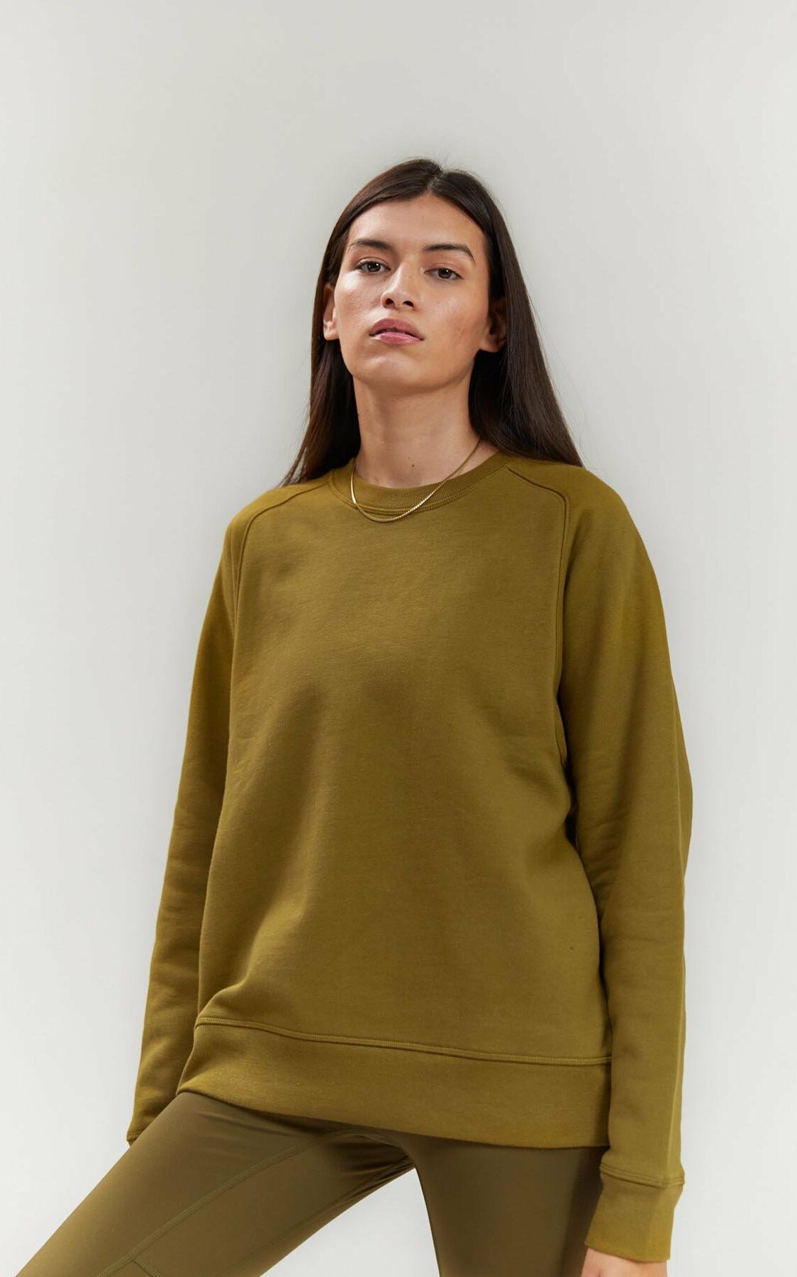 A white person with long straight brown hair stands in an olive colored crewneck.-What does Androgyny in Plus Size Fashion Mean to You? Part I