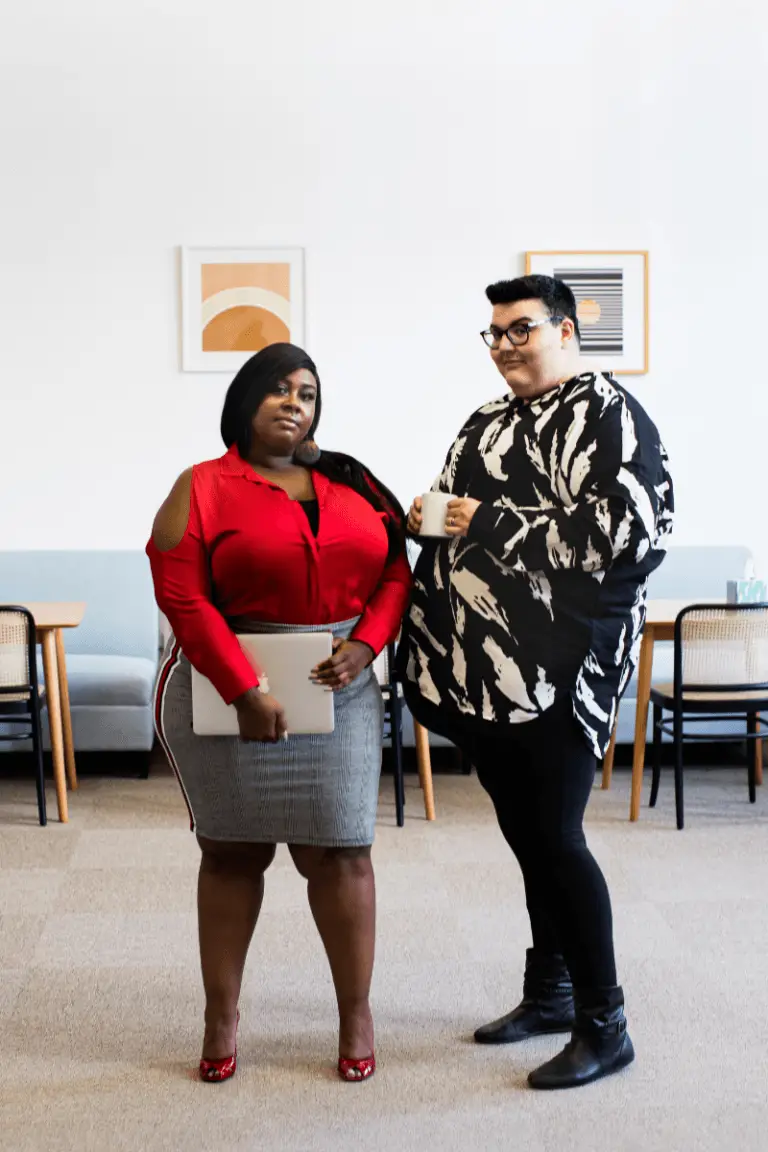 Two people stand in an office waiting room. On the left is a Black person with short cropped straight hair wearing a red v-neck long sleeve shirt with a grey skirt. Next to them stand a slightly taller white person with black hair wearing an oversized black and white designed long-sleeve shirt over black skinny jeans.