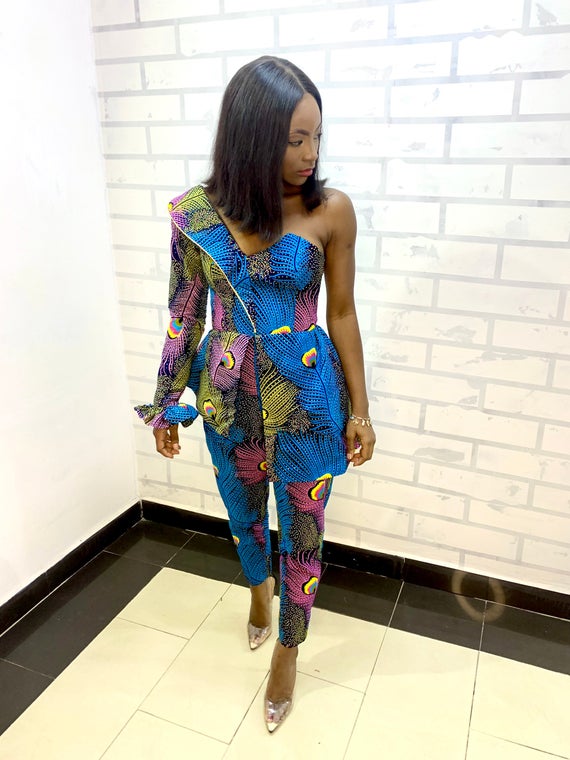 a Black person stands in a custom jumpsuit. the jumpsuit is brightly patterned in blues and pinks and has one sleeve, while the other shoulder is sleeveless. the jumpsuit has tightly cropped pants and a corset shape.