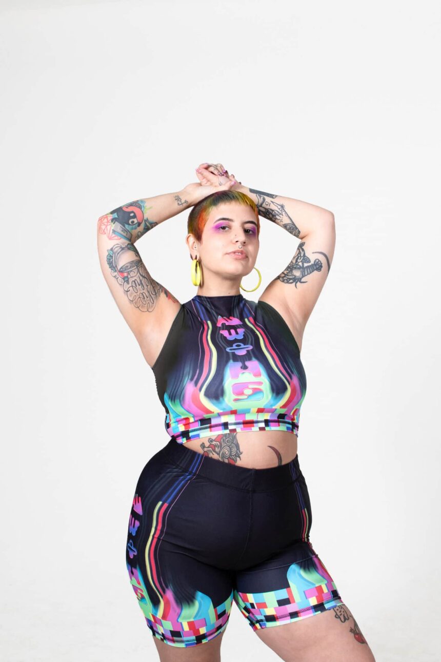 A white person with short, colorful hair stands with their arms over their head. they have a few tattoos on their arms and stomach. they are wearing a neon and colorful crop top by MW+HS with high waisted biker shorts.