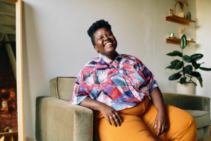 Lydia Okello sits on a chair, in orange pants and a colorful button up top, they are smiling