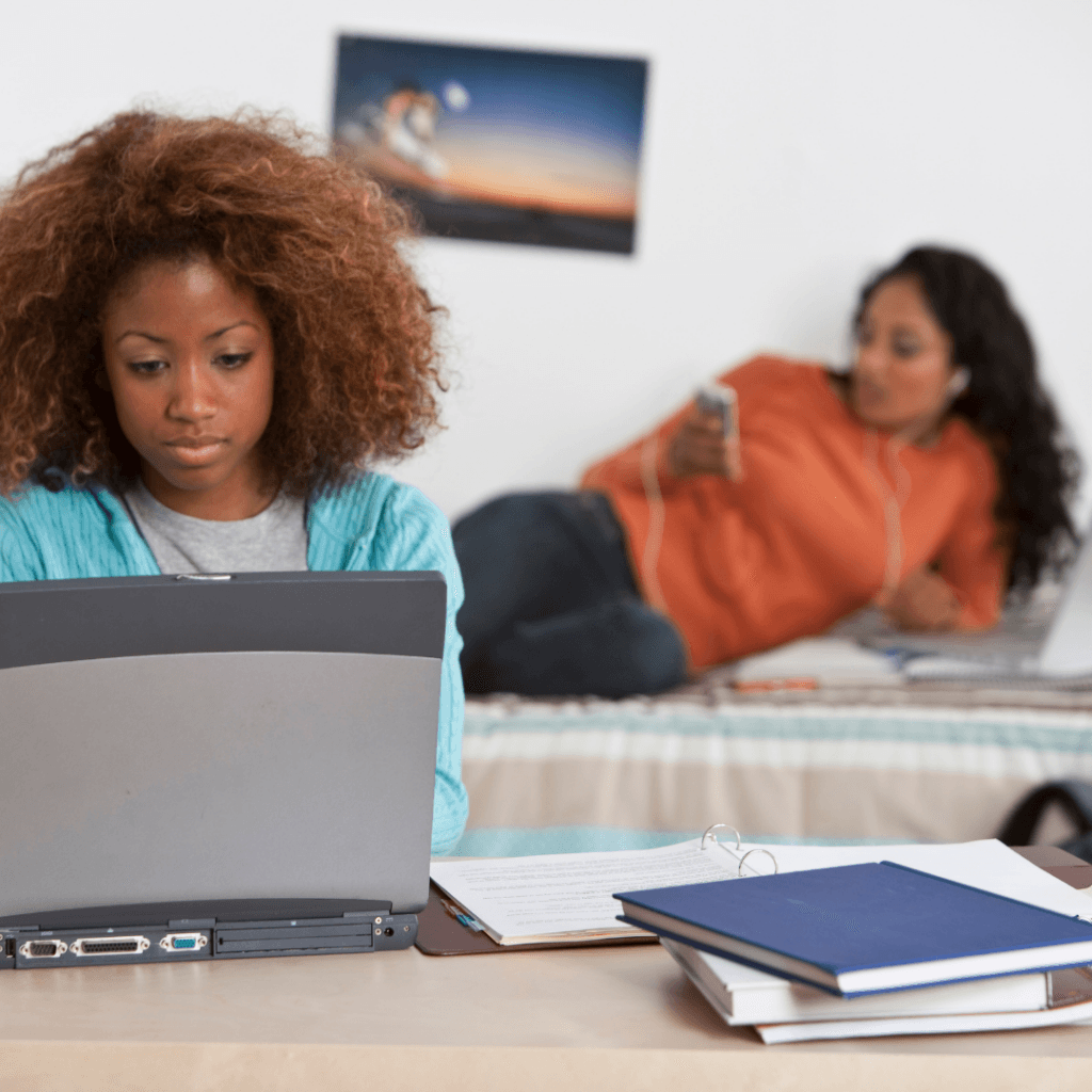 Two college students in a room, one on the bed and one on her computer