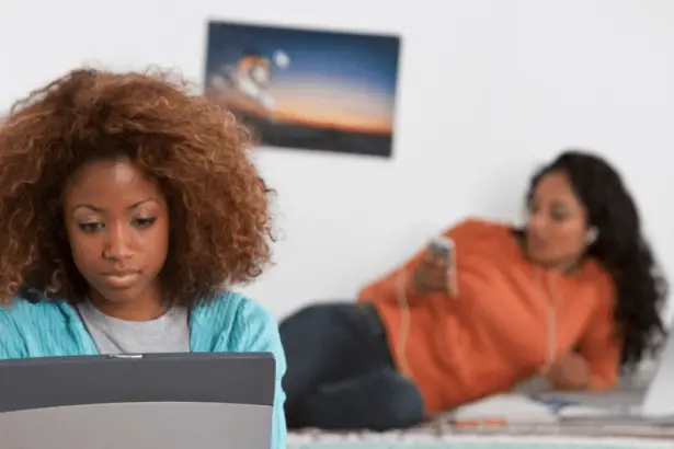 Two college students in a room, one on the bed and one on her computer