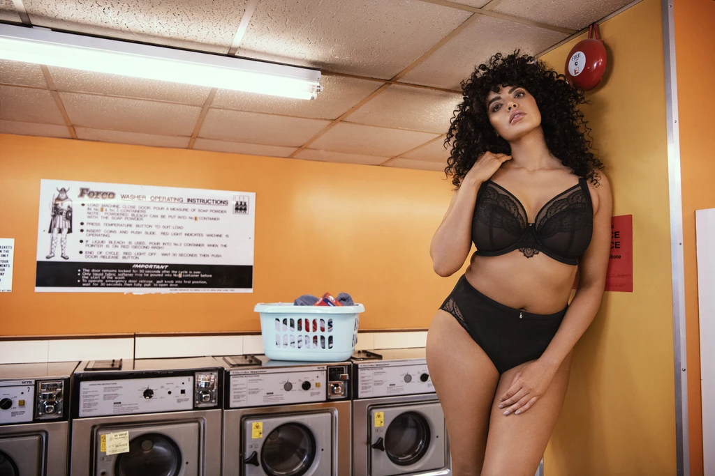 Here are 5 Full Figured Bra Brands to KNOW!Image of a woman standing in a laundromat setting wearing the Priya plunge bra