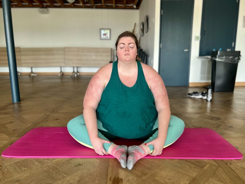white plus size woman with hair braided back, matching teal yoga set, sitting on a yoga mat in meditation