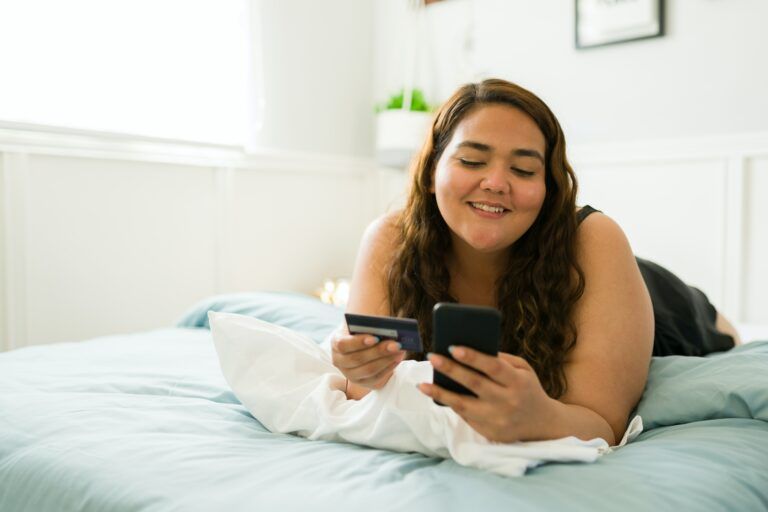 plus size light skinned woman with shoulder length brown hair looking at a phone holding a debit card