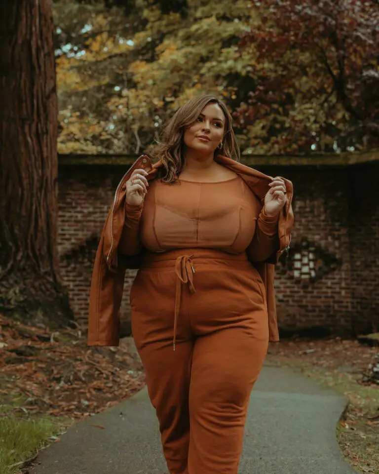 Brown haired white woman in orange pants and top with jacket shrugged around her shoulders smirking