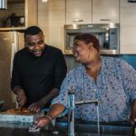 a black woman with short hair and a black man with a fade haircut smiling at each other while cutting vegetables in the kitchen