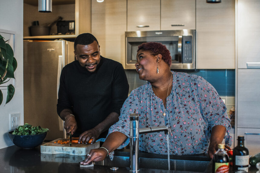 a black woman with short hair and a black man with a fade haircut smiling at each other while cutting vegetables in the kitchen