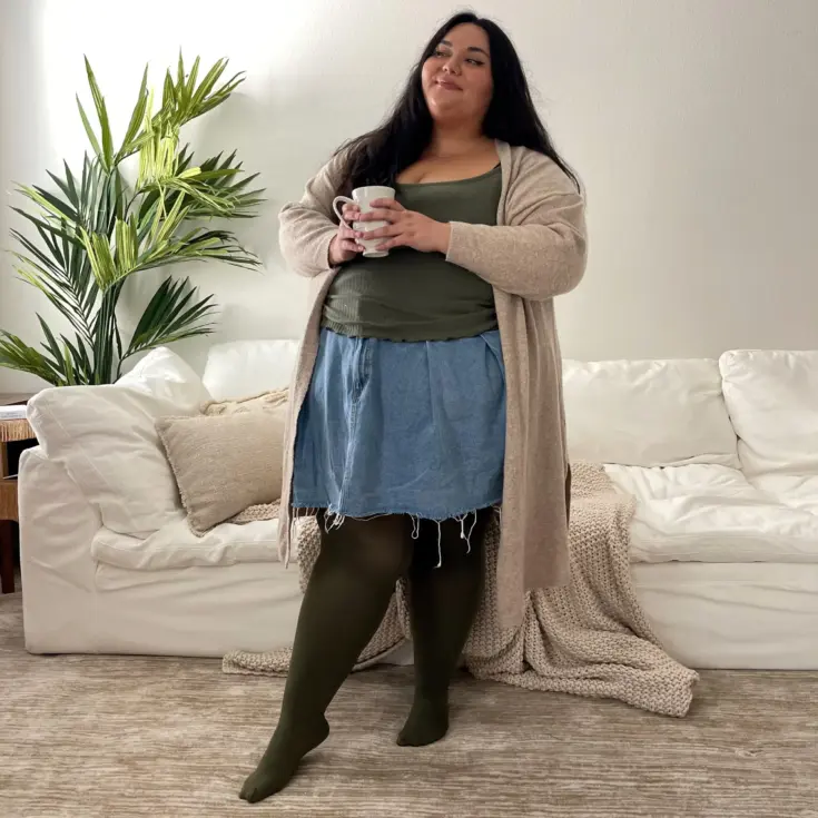 light skinned latina in a neutral decorated room wearing a denim skirt, tights, and oversized cardigan smiling.