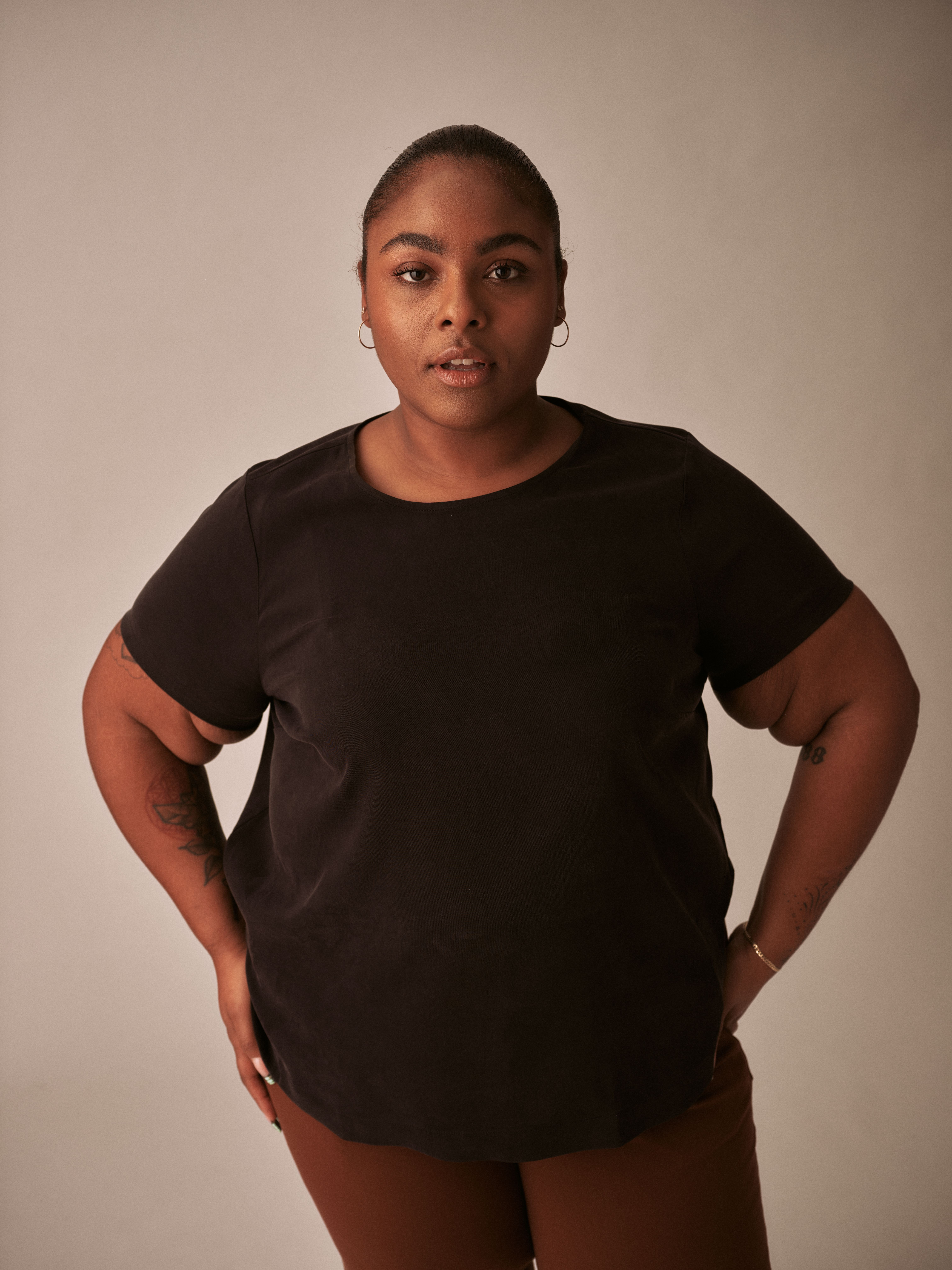 Black feminine presenting person with hair slicked back in black tea shirt looking into camera
