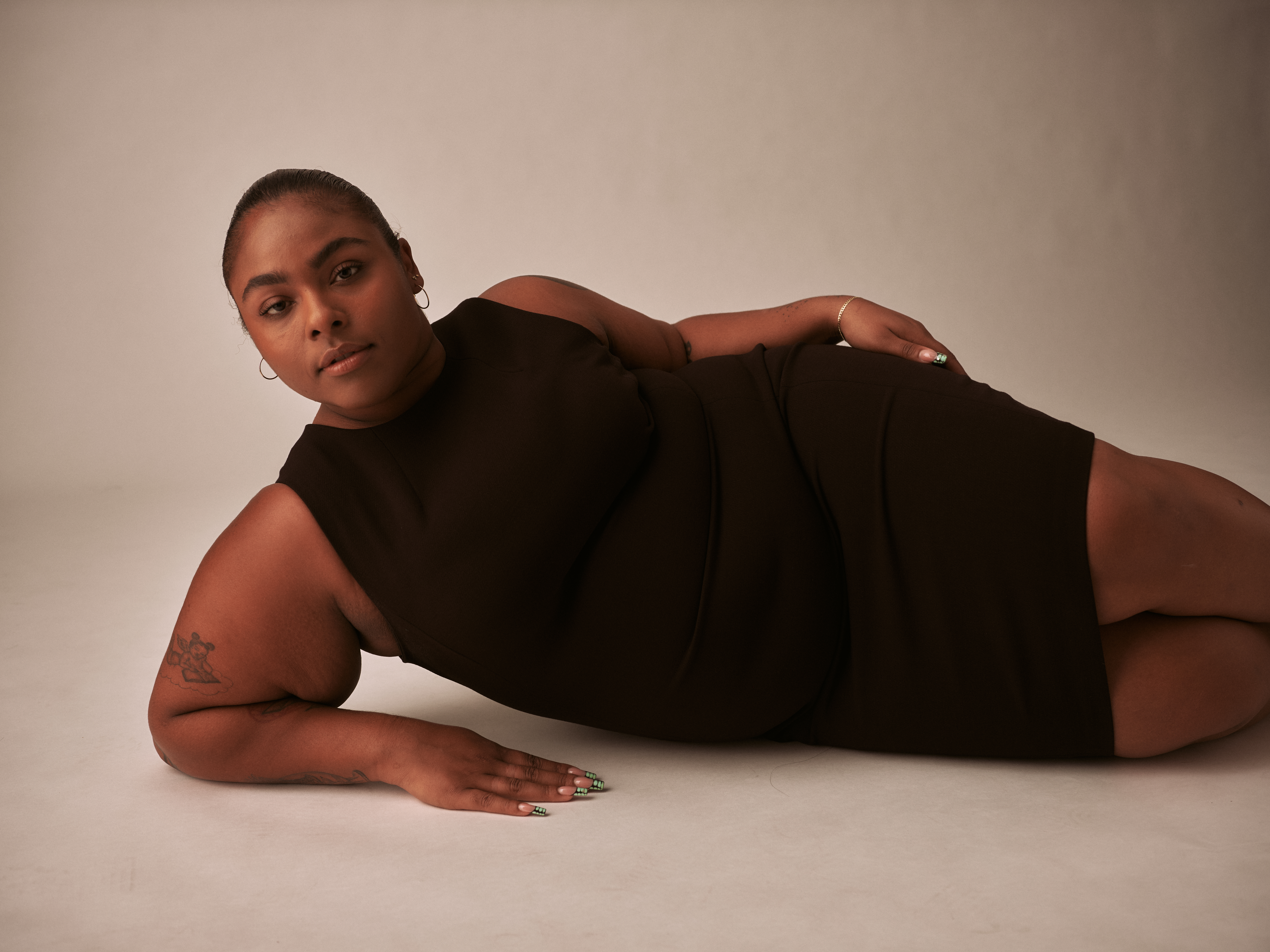 black feminine presenting person laying on side in dress propped up on elbow looking into camera.