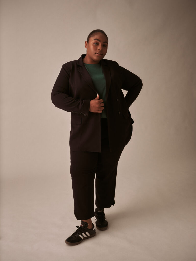 black feminine presenting person wearing a black suit with a green top posed with hand on hip looking into camera with sass.