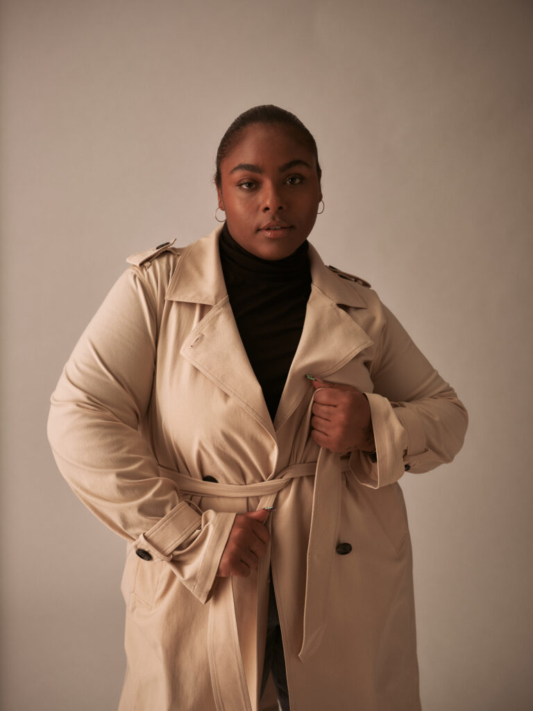 black feminine presenting person  wearing trench coat, pulling wrap belt tight looking into camera smirking.