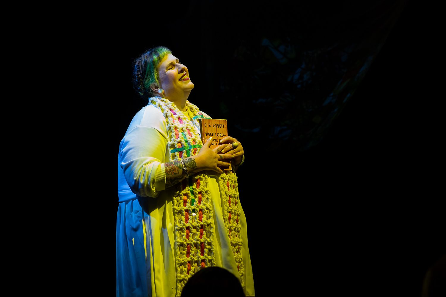 a white plus sized person with black and teal hair wearing a stole weaved with measuring tape and rope holding a book looking up smiling with eyes closed