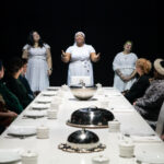 Brown skinned feminine looking person standing at the head of a full table with arms outstretched forward