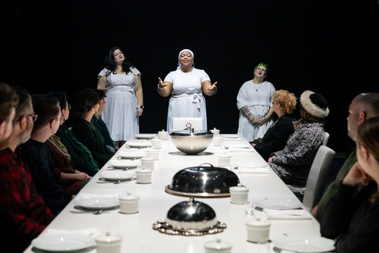 Brown skinned feminine looking person standing at the head of a full table with arms outstretched forward