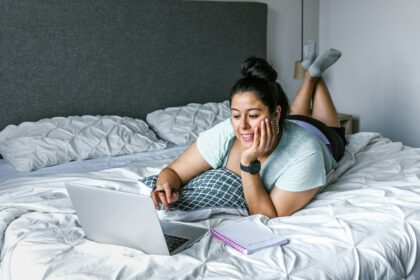 light skinned woman with dark hair piled in a bun on head laying on bed browsing on laptop smiling