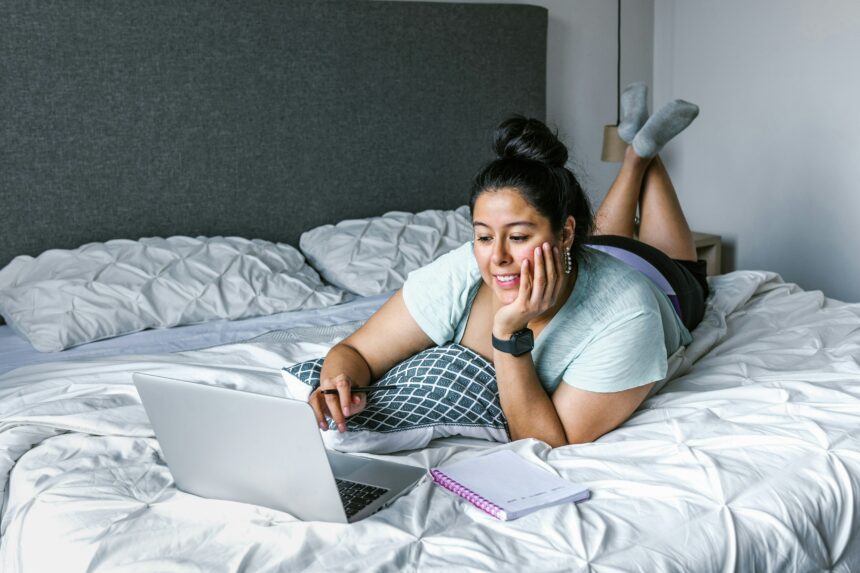 light skinned woman with dark hair piled in a bun on head laying on bed browsing on laptop smiling