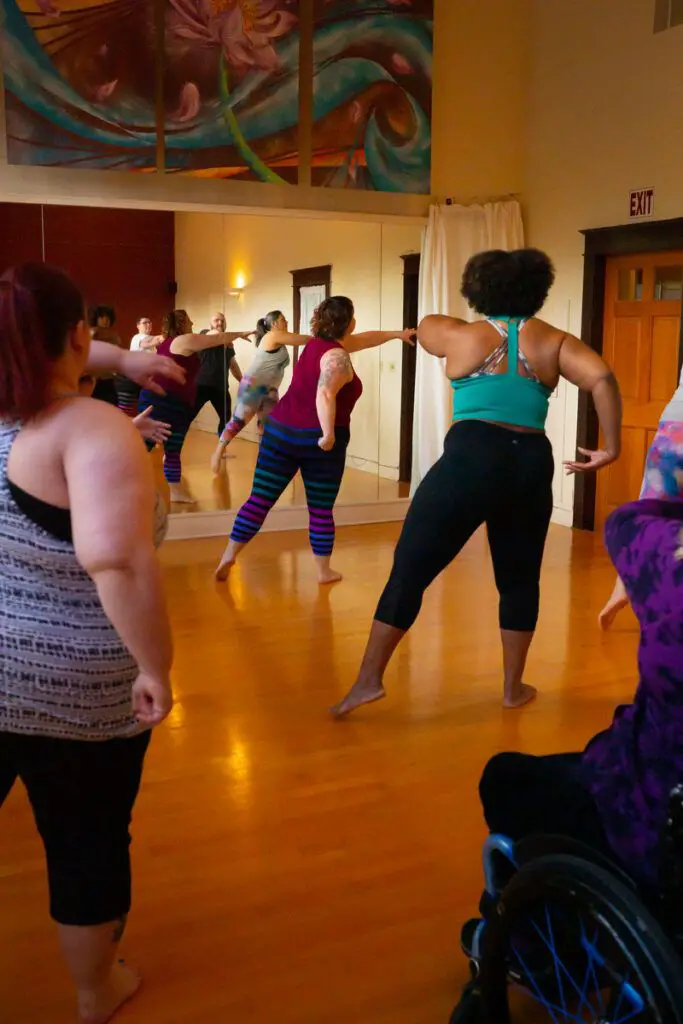 Students move, stretch, and dance in a weight-inclusive fitness dance class. The class includes people who are plus size or fat, people of color, and people with visible disabilities using mobility aids.