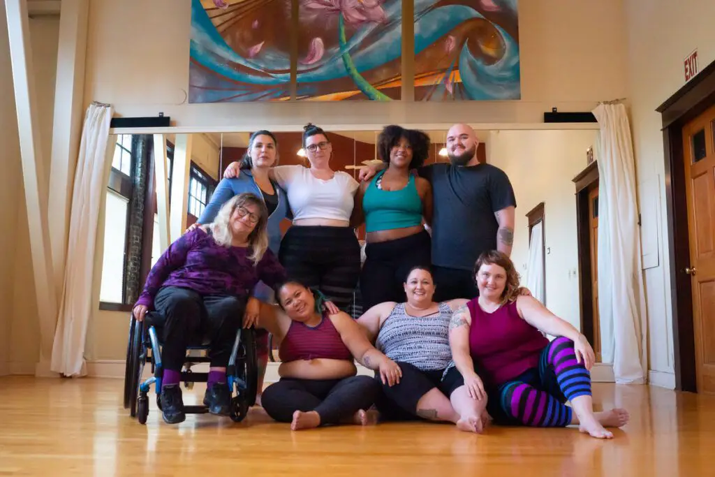 A diverse group of people participates in a weight-inclusive fitness class in a yoga studio with wooden floors, red walls, and mirrors.