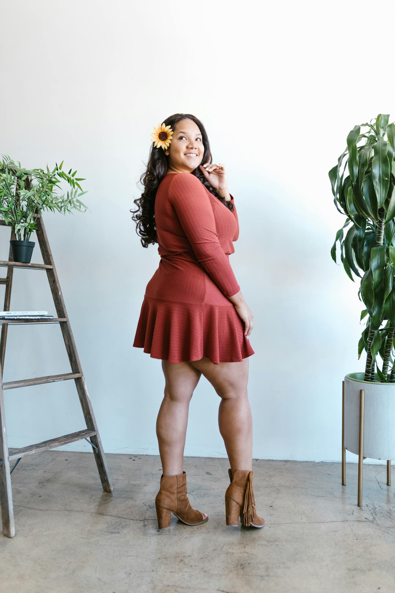 A confident plus-size woman in a fashionable dress smiles gracefully in a studio setting with indoor plants.