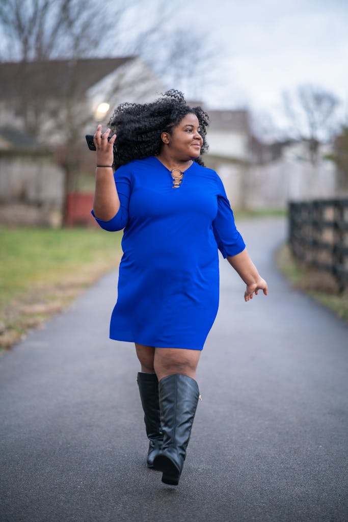 A confident woman in a blue dress walks with a smile on a suburban street.