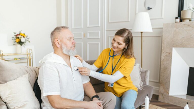 A nurse uses a stethoscope for a home check-up on a senior adult in a cozy living room.