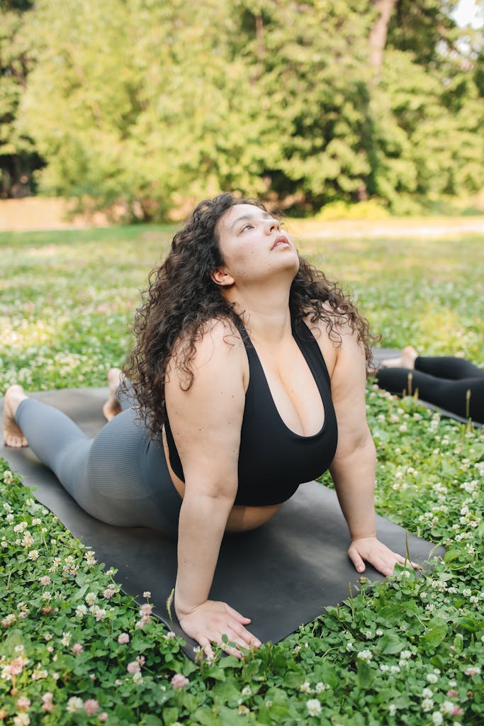 A plus size woman doing yoga on a mat outdoors in a green park setting.