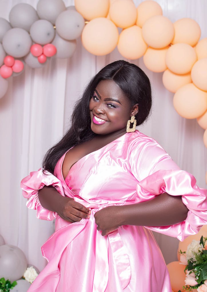 A smiling plus size woman in a pink dress poses elegantly with balloons in the background.
