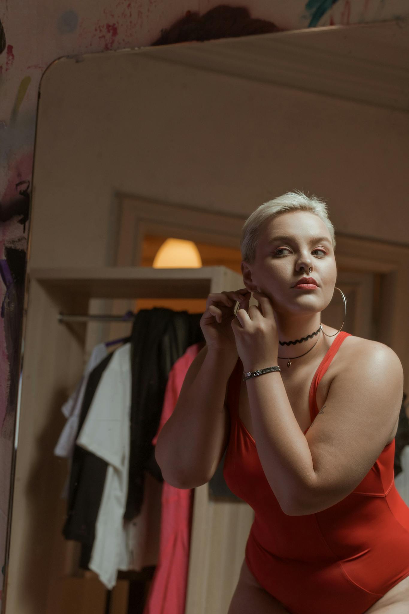 A woman in a red bodysuit adjusts hoop earrings while looking in the mirror indoors. Plus size accessories