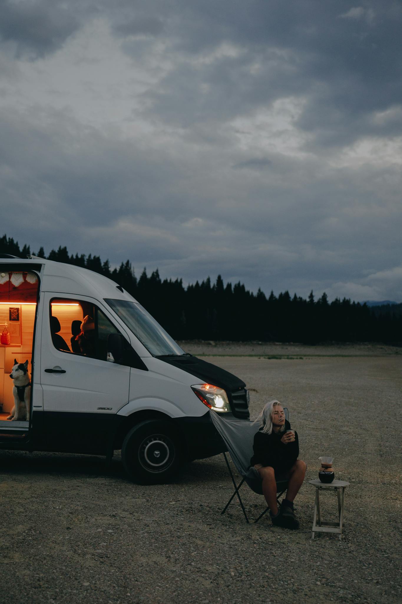 An adult woman relaxes outside a campervan, sitting under cloudy skies with her dog by her side.