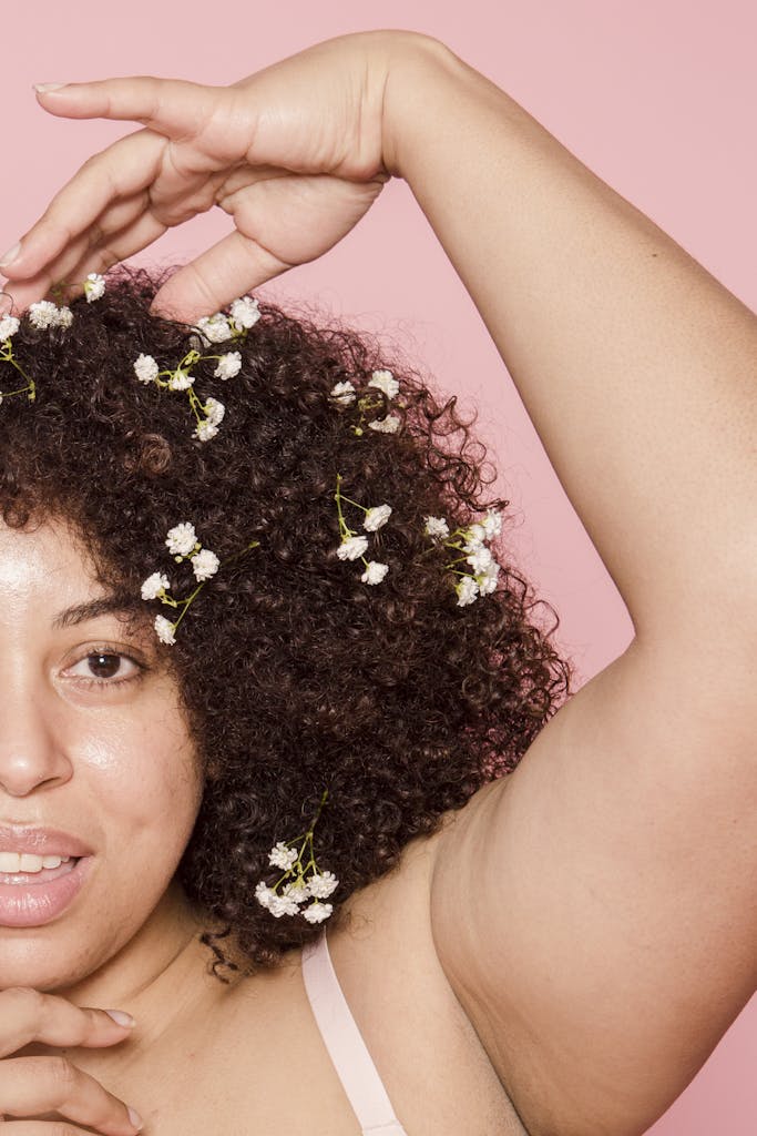 Crop content overweight African American female model with creative small flowers in black hair looking at camera with raised arm on pink background