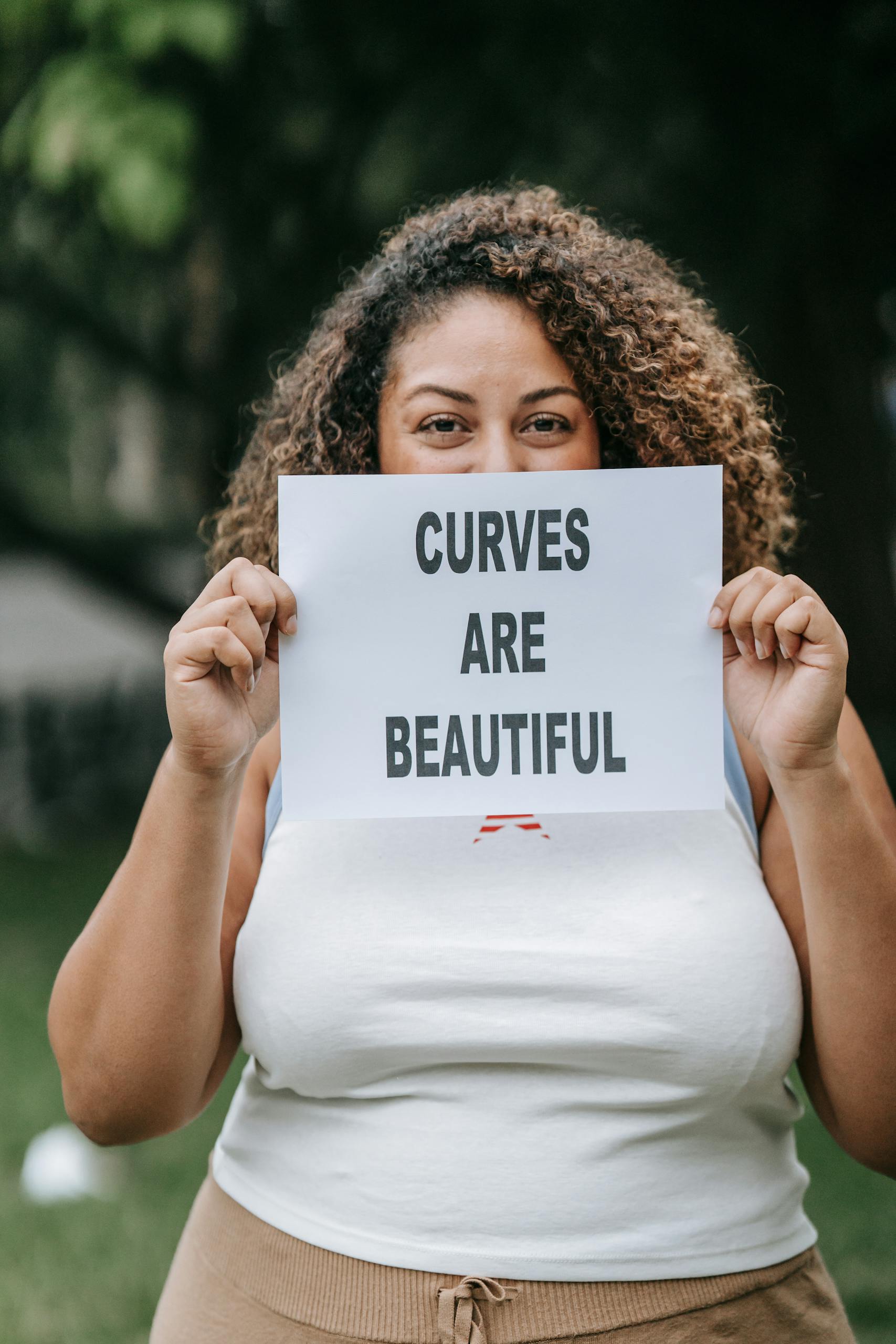 Curvy woman proudly holding a 'Curves are Beautiful' sign outdoors, promoting self-love and body positivity.