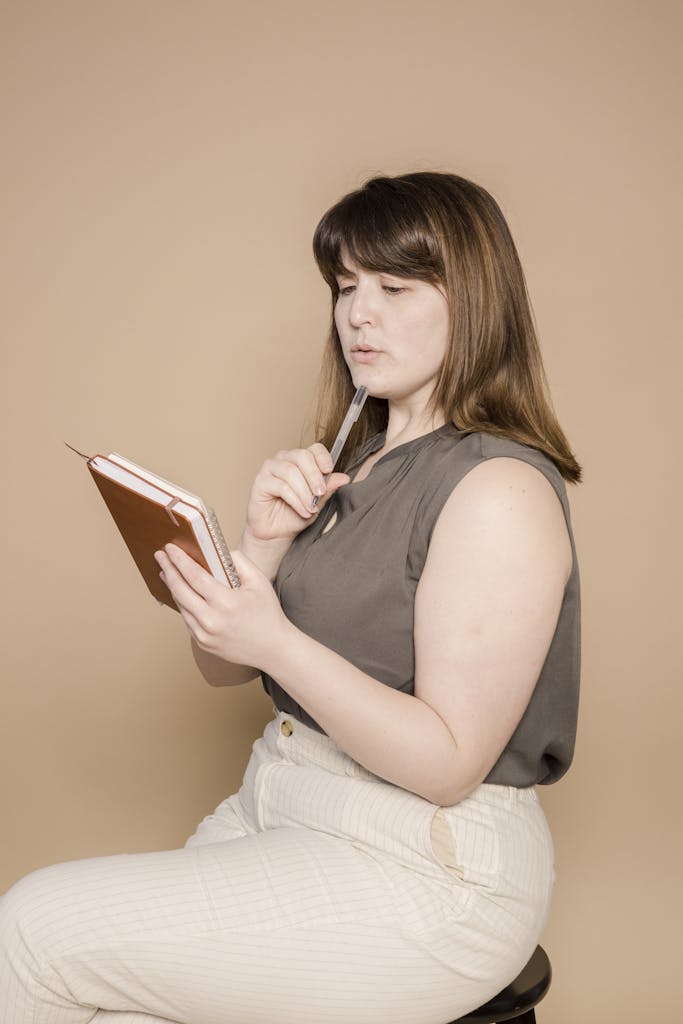 Plus-size woman pensively writing in a notebook against a beige backdrop in a studio shoot.