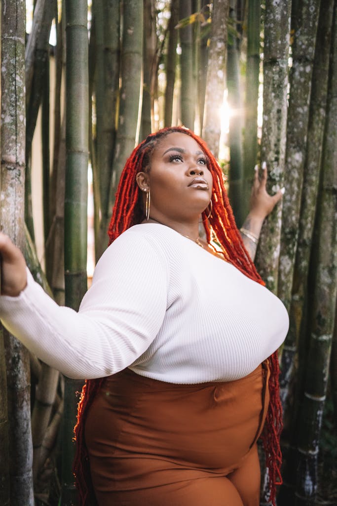 Portrait of a confident woman with red hair standing among tall bamboo under sunlight.
