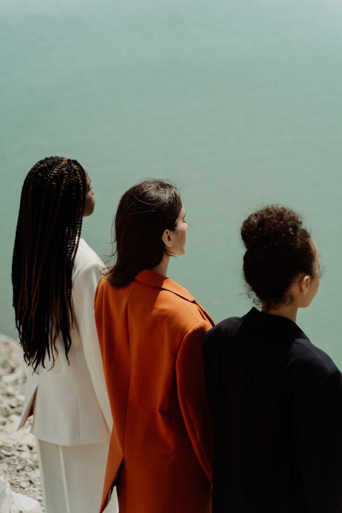 Three women stand by water, dressed elegantly in vibrant fashion, showcasing diversity.