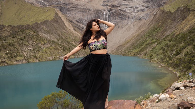 Woman in a flowing skirt poses by a turquoise mountain lake and rocky hills.