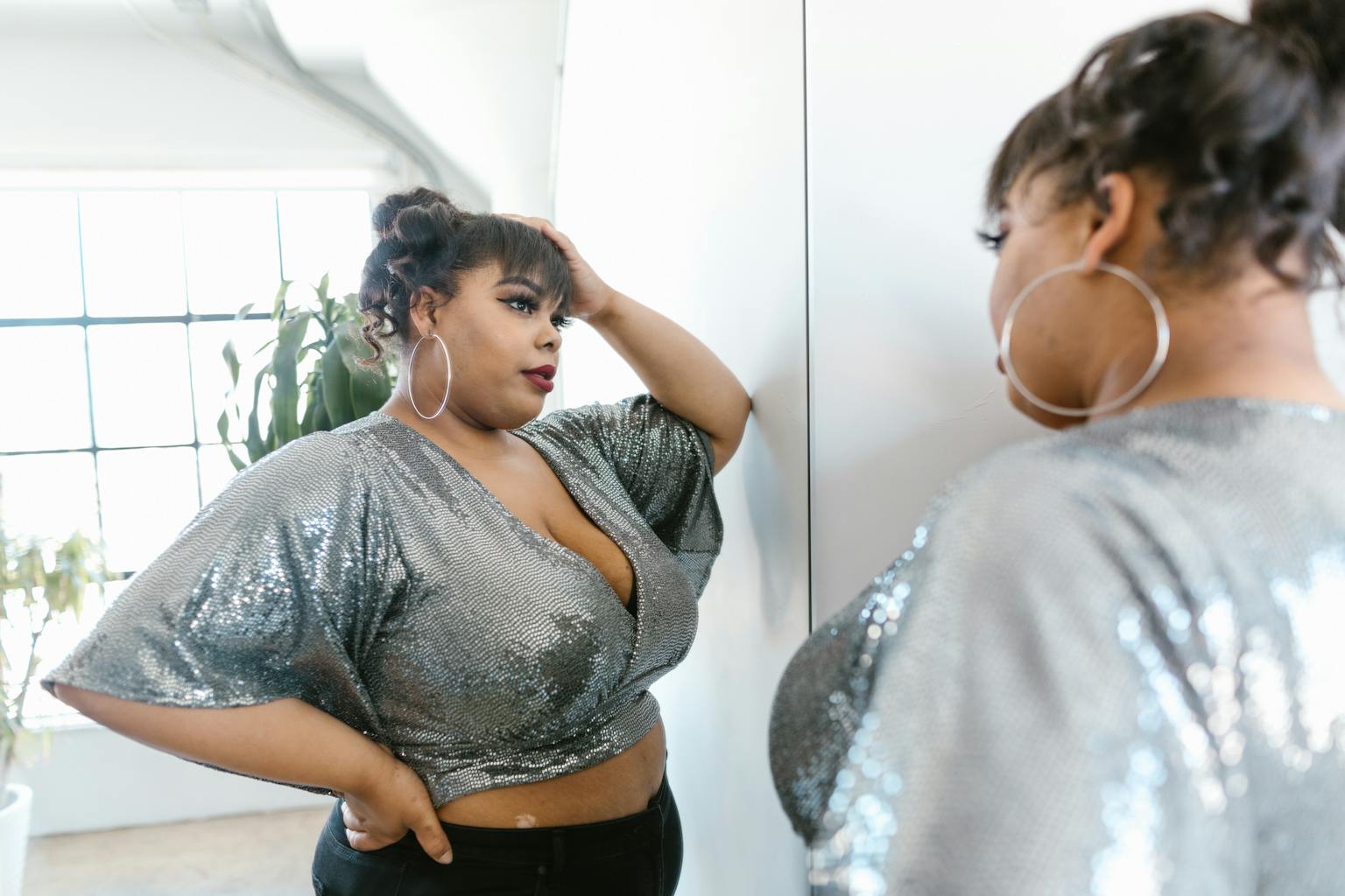 A confident plus size woman in a sparkling top poses before a mirror, embracing self love in style.