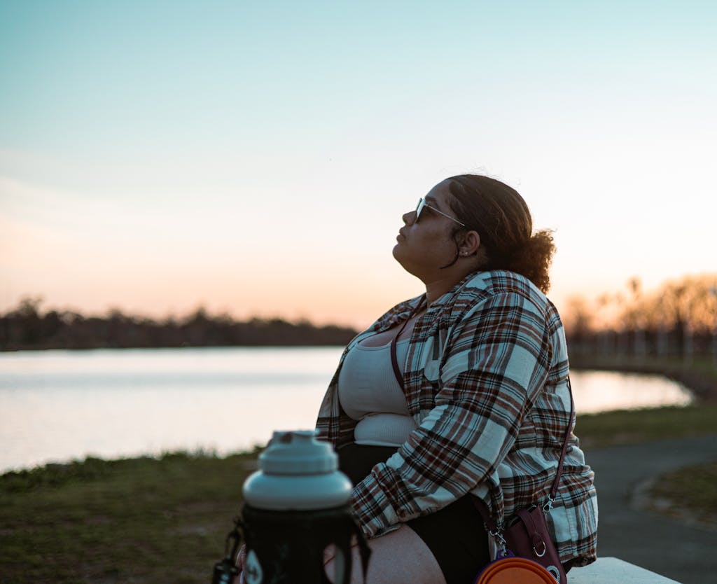 A fashionable woman in plaid relaxes by the lakeside at sunset, wearing sunglasses and looking serene.