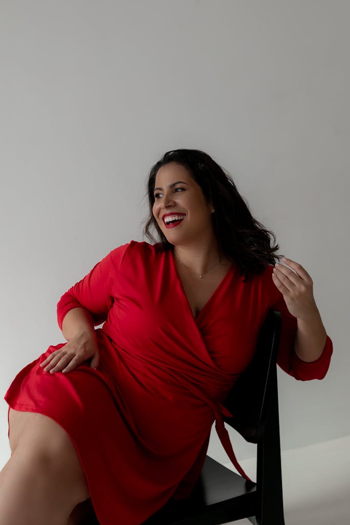 A joyful woman in a striking red dress sits on a chair, exuding elegance and confidence.

plus size confidence in 2026