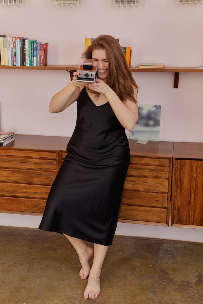 A smiling woman in a black dress takes a photo with a vintage Polaroid camera indoors.